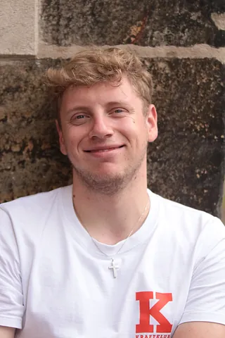 Jakob Schmidtke mit lockigem, blondem Haar trägt weißes T-Shirt mit rotem K auf der Brust und silbernes Kreuz als Halskette, vor grober Steinmauer stehend.