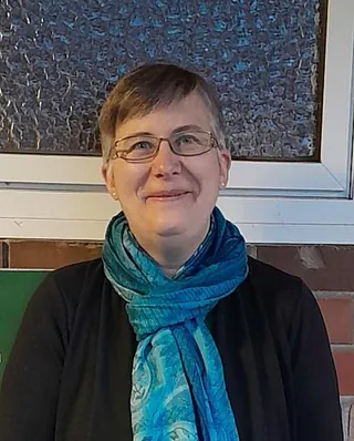 Dorothee Heinz mit kurzem Haar trägt schwarzen Pullover und türkisfarbenen Schal, sitzt vor einer Wand mit strukturierter Glasfläche und Ziegelsteinen.