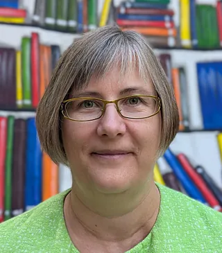 Dorothee Heinz mit kurzem Haar trägt schwarzen Pullover und türkisfarbenen Schal, sitzt vor einer Wand mit strukturierter Glasfläche und Ziegelsteinen.