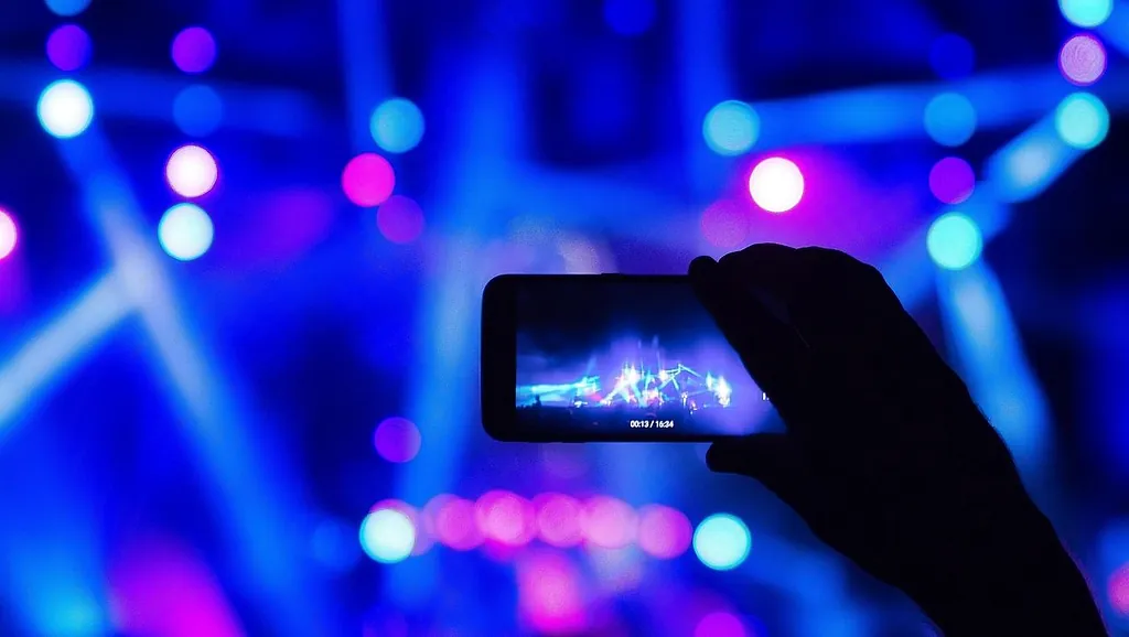 Hand hält Smartphone, das eine Bühne mit bunten Lichtstrahlen bei einer Veranstaltung zeigt