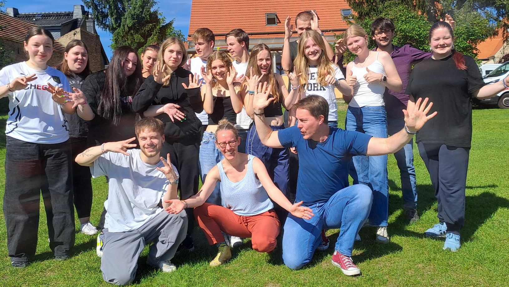 Eine Gruppe Konfirmand:innen steht auf einer Wiese vor Wohnhäusern, einige hocken, andere stehen, alle zeigen mit Händen verschiedene Gesten.