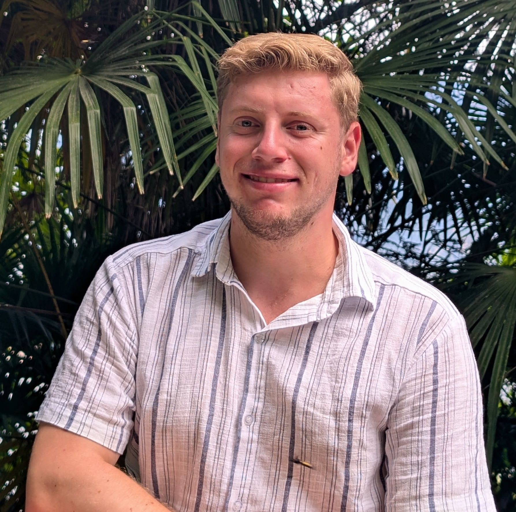 Jugendmitarbeiter Jakob Schmidtke trägt ein kurzärmeliges, weißes Hemd mit grauen Streifen und steht vor dichtem, grünem Palmenlaub.
