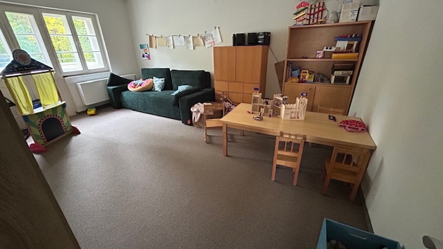 Spielzimmer Gemeindehaus mit Sofa, Holztisch und Stühlen, Spielzeugburg, Regalen und Fenster.