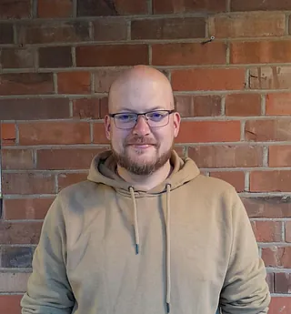 Mathias Hugo trägt einen hellbraunen Kapuzenpullover vor einer roten Backsteinwand.