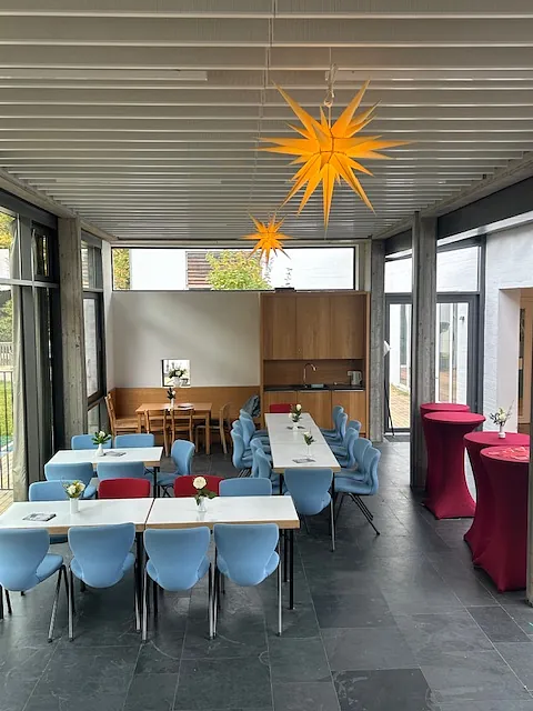Offenes Cafe am Dienstag mit blauen Stühlen, weißen Tischen, roten Stehtischen und gelben Sternlampen an der Decke.