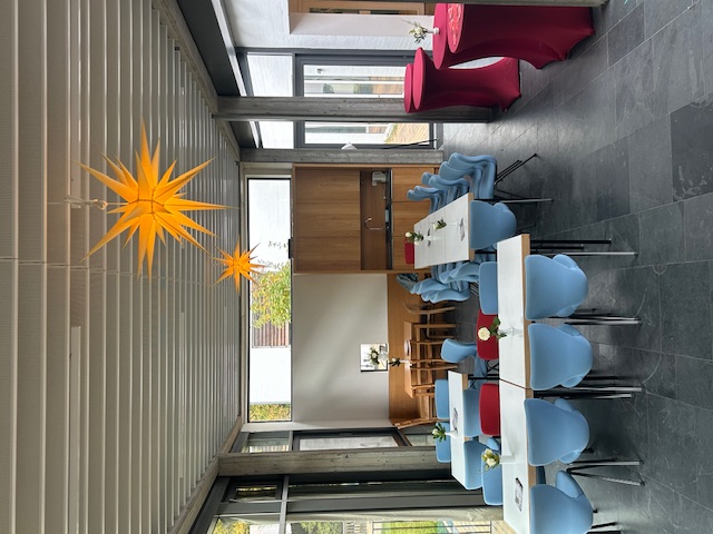 Offenes Cafe am Dienstag mit blauen Stühlen, weißen Tischen, roten Stehtischen und gelben Sternlampen an der Decke.
