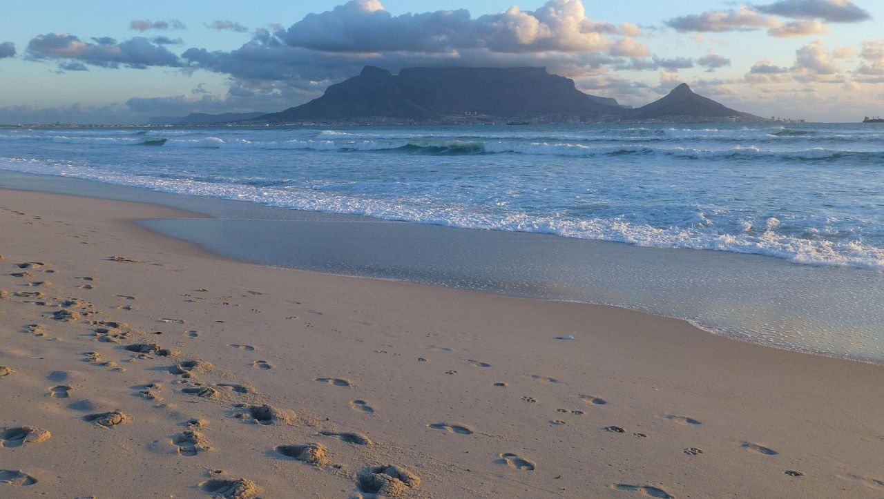 Sandstrand mit Fußspuren, dahinter Meer mit Wellen und im Hintergrund der Tafelberg unter bewölktem Himmel