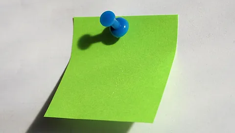 Grüner Notizzettel mit blauem Reißnagel an weißer Fläche befestigt, Schatten sichtbar