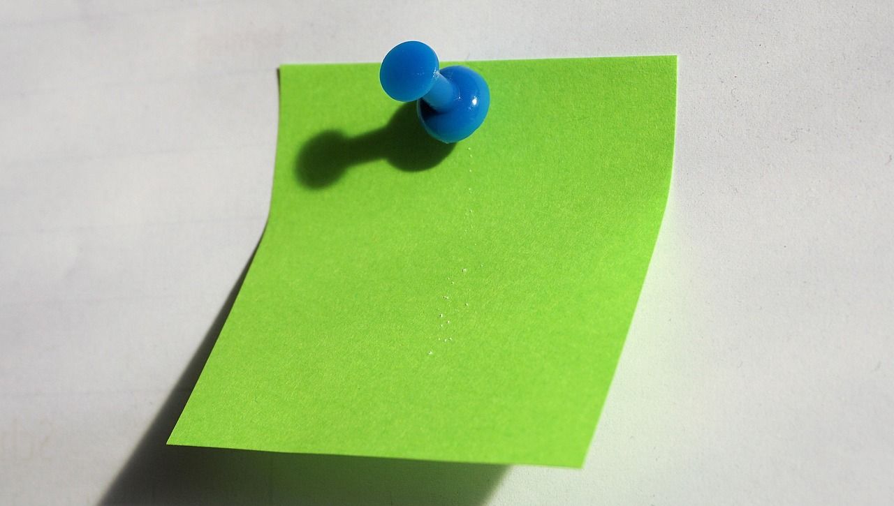 Grüner Notizzettel mit blauem Reißnagel an weißer Fläche befestigt, Schatten sichtbar