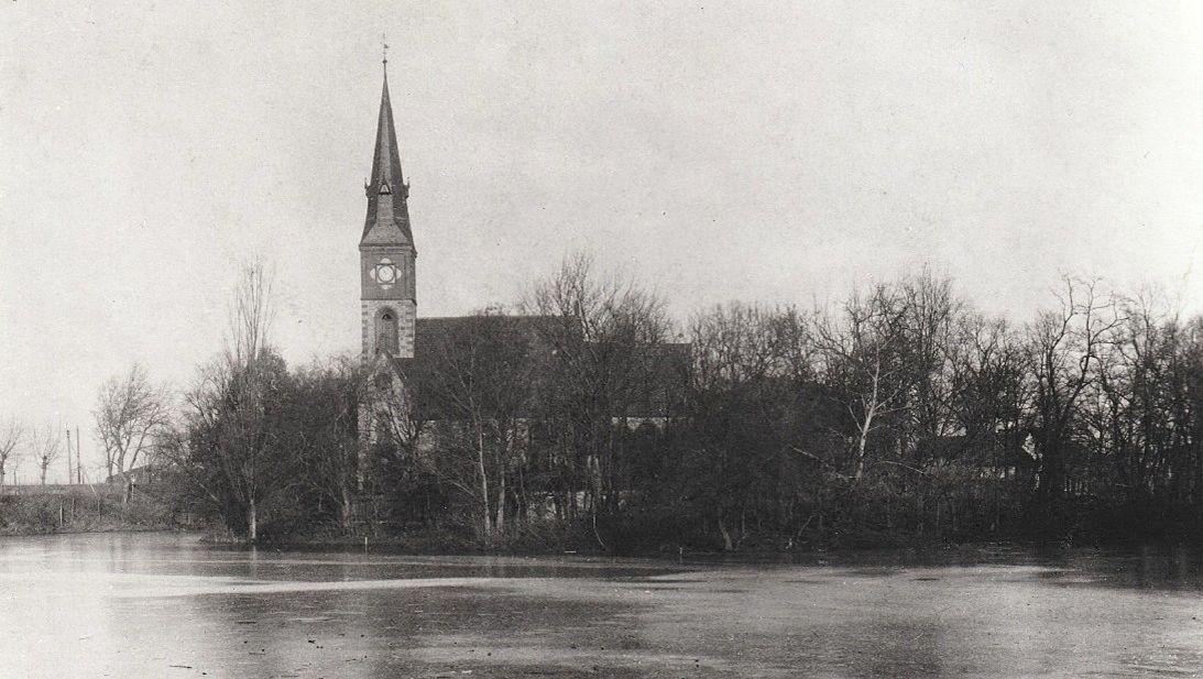 Kirche Britz Dorf mit spitzem Turm und Uhr, umgeben von kahlen Bäumen, im Vordergrund ein zugefrorener See