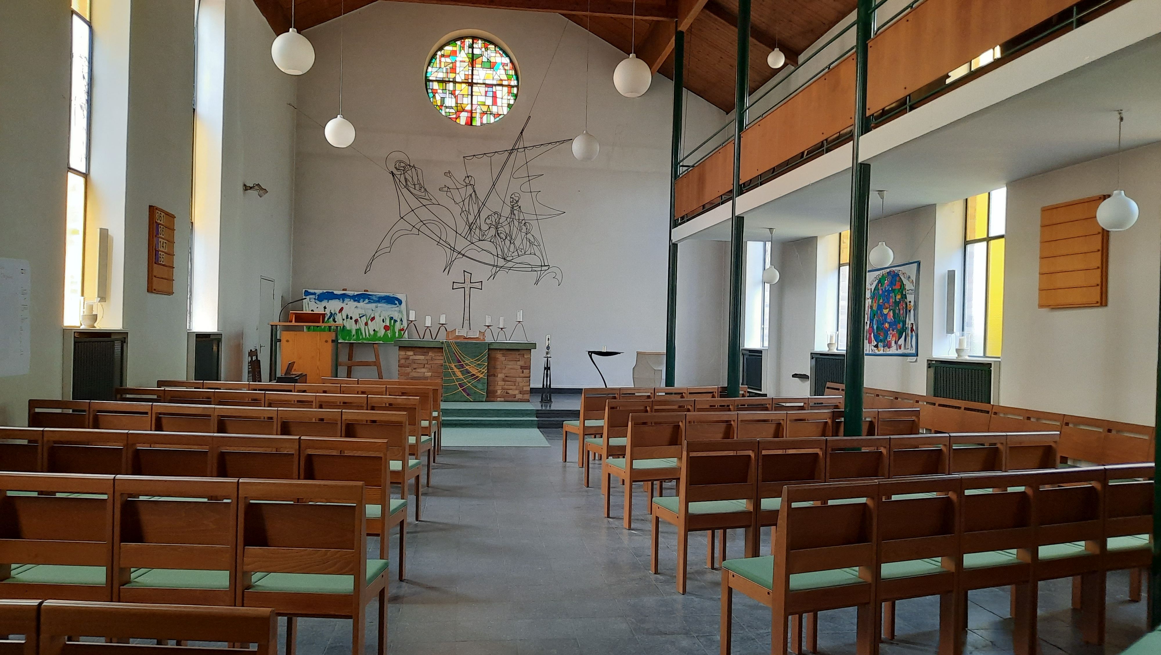 Kirchraum Hephatha mit Holzbänken, Altar, rundem buntem Fenster und schwarzer Linienzeichnung an der Wand unter Holzdecke mit weißen Kugelleuchten.