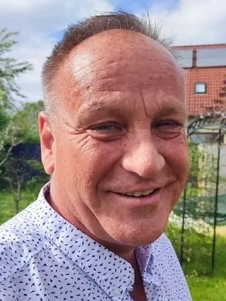 Andreas Seide trägt ein weißes Hemd mit schwarzem Muster, steht im Garten vor einem Haus mit rotem Dach und Fenstern.